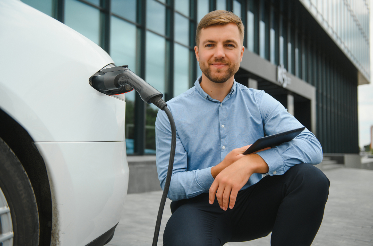 Man in een blauwe blouse knielt naast een elektrische auto die wordt opgeladen, met een tablet in zijn hand.