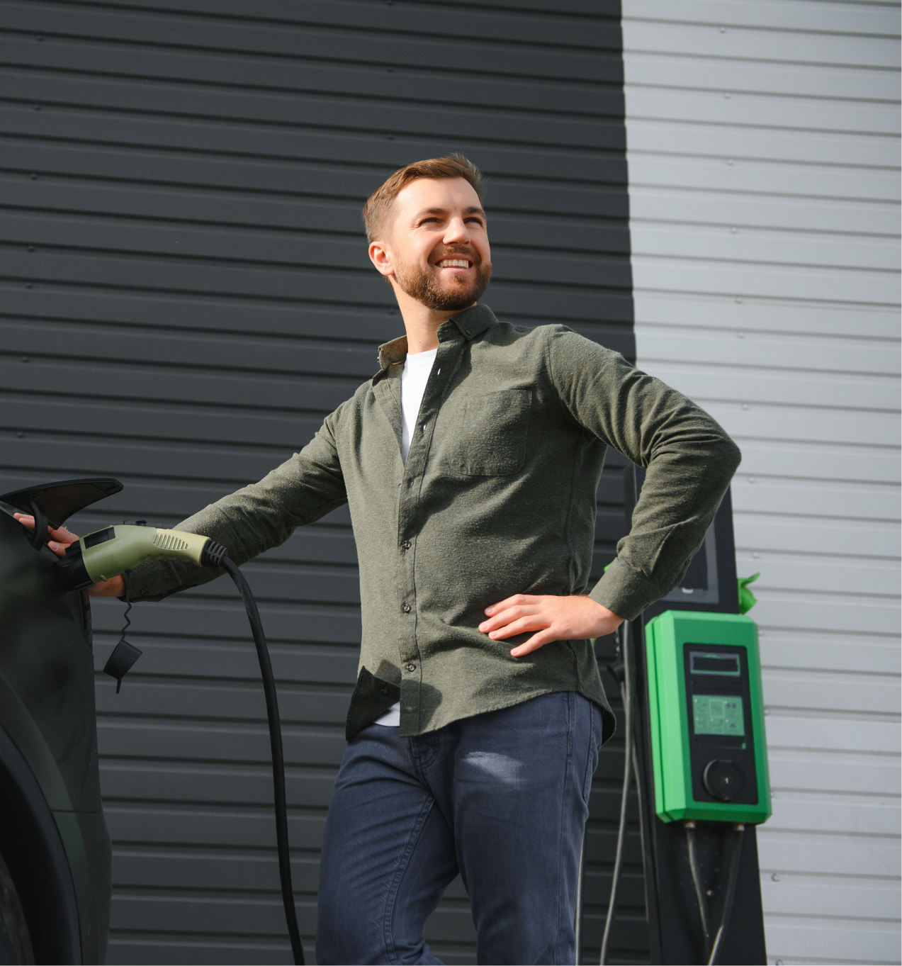 Man in een groen overhemd staat met een opgeluchte glimlach bij een elektrische auto terwijl hij de laadkabel vasthoudt.