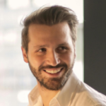 Man met kort bruin haar en een glimlach, gekleed in een witte blouse, poseert voor een lichtgevend achtergrond.