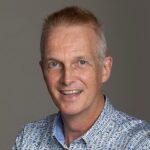 Martien poseert lachend in een blauw-wit patroonoverhemd voor een neutrale achtergrond.