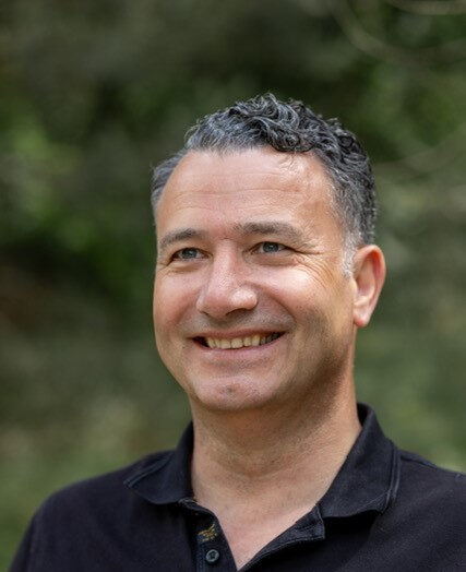 Man met kort krullend haar en zwarte polo poseert lachend buiten voor een groene achtergrond.