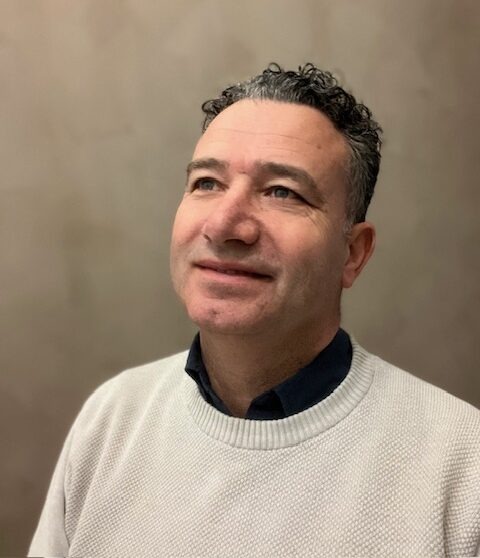 Man met kort krullend haar en zwarte polo poseert lachend buiten voor een groene achtergrond.