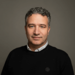 Man met kort krullend haar en zwarte trui poseert glimlachend voor een grijze achtergrond.