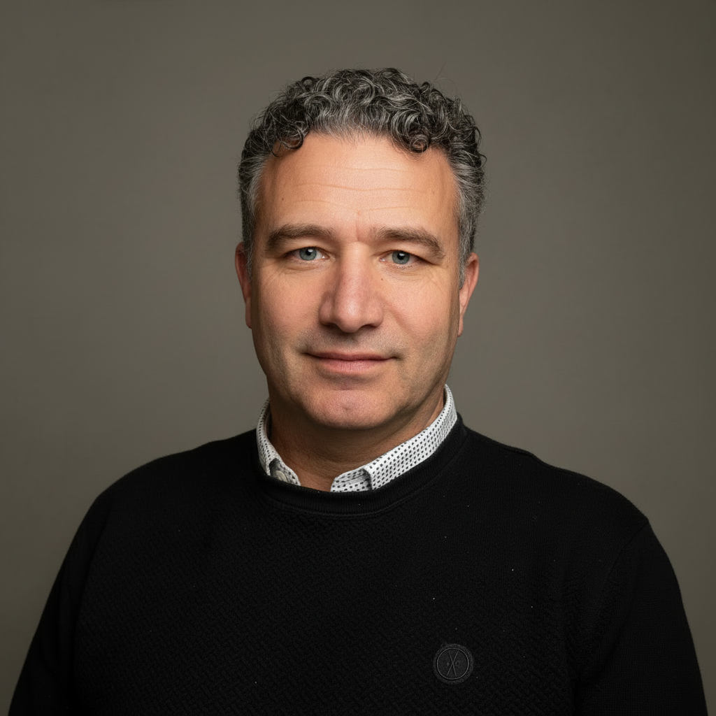 Man met kort krullend haar en zwarte trui poseert glimlachend voor een grijze achtergrond.