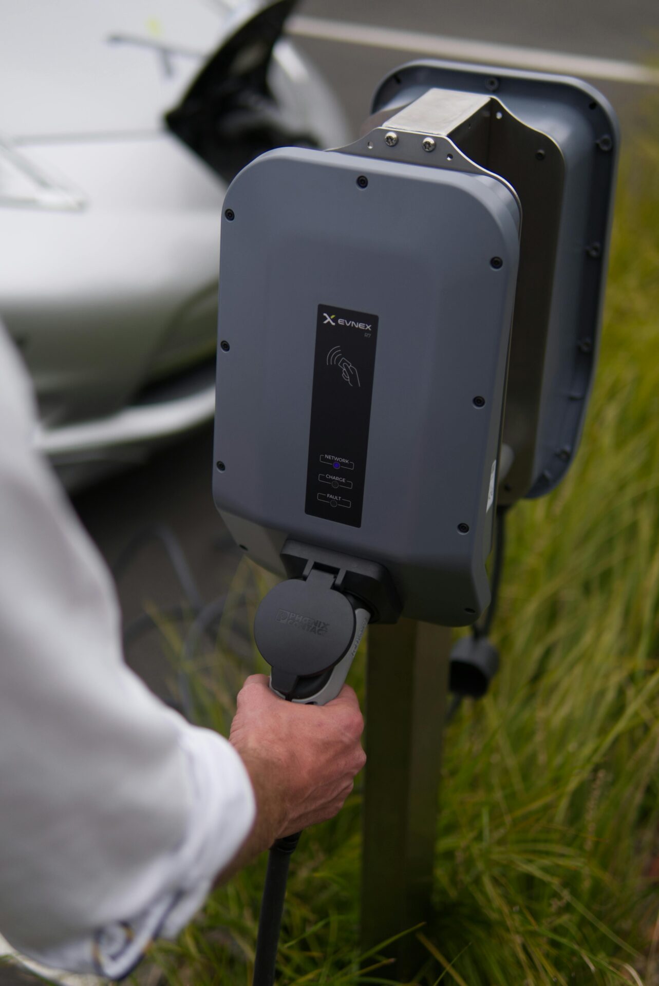 Close-up van een persoon die een laadkabel inplugt in een grijs EV-laadstation van het merk EVNEX, met gras op de achtergrond en een zilverkleurige elektrische auto gedeeltelijk zichtbaar links in beeld.