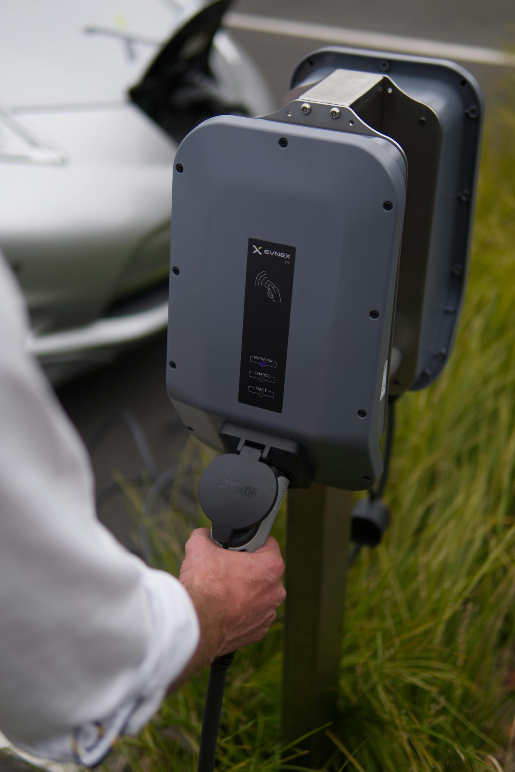 Close-up van een persoon die een laadkabel inplugt in een grijs EV-laadstation van het merk EVNEX, met gras op de achtergrond en een zilverkleurige elektrische auto gedeeltelijk zichtbaar links in beeld.