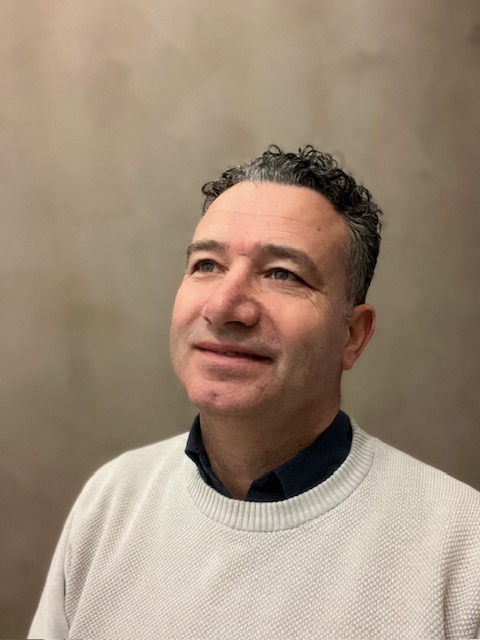 Man met kort krullend haar en zwarte polo poseert lachend buiten voor een groene achtergrond.